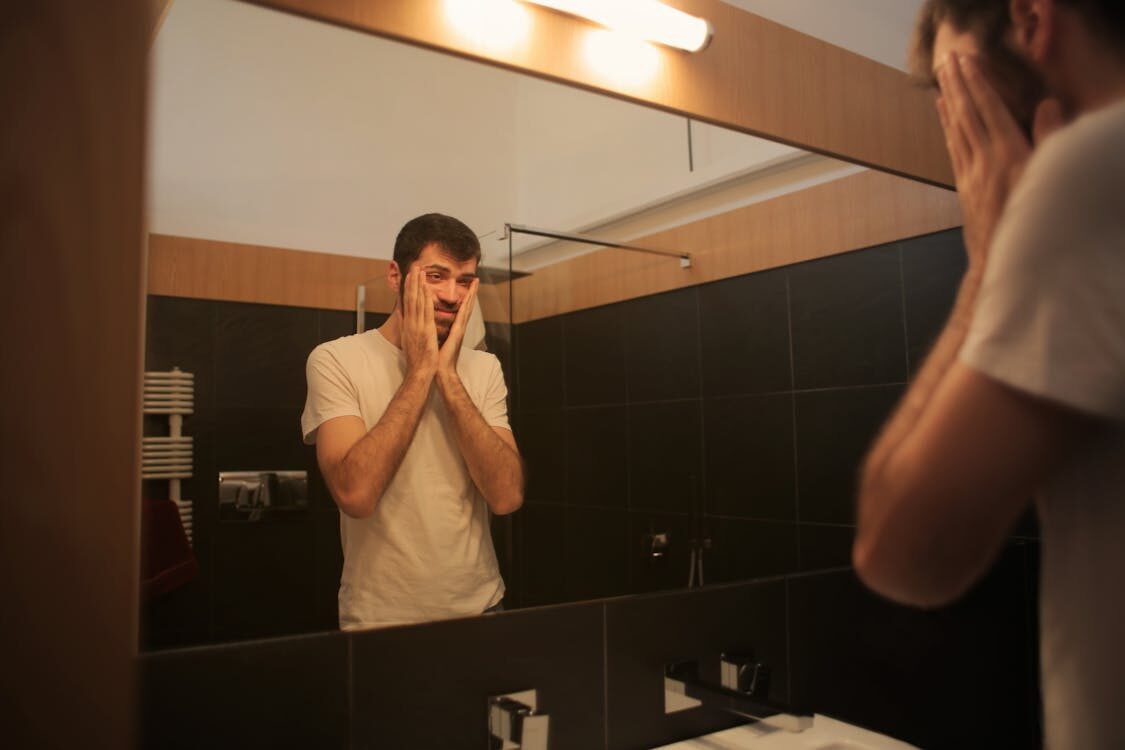 An Individual Needing Anti-Aging for Men Manually Tightens His Facial Skin While Looking at His Reflection in a Bathroom Mirror