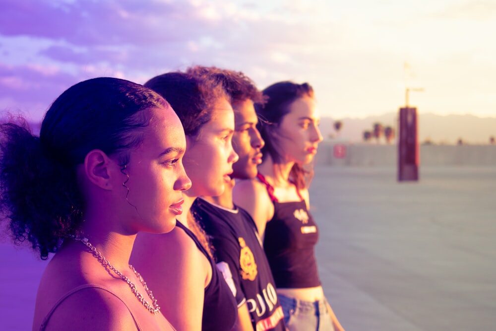 Profile view of four teenagers lined up in a gradient of skin tones under a purple-hued sunset sky.