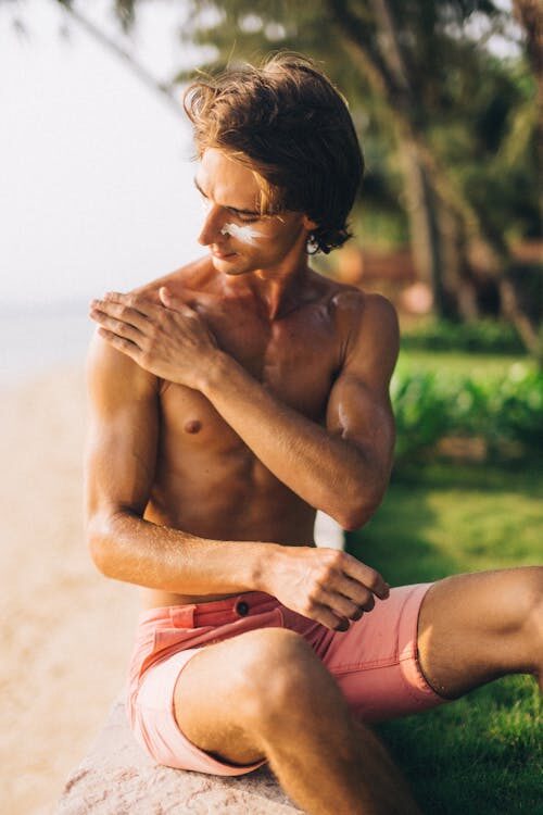 A male in pink shorts applying sunscreen on his face and shoulders. 